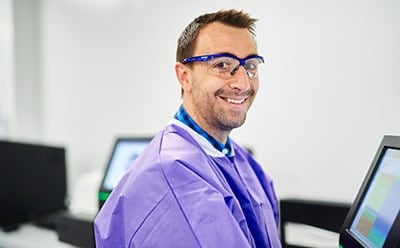 Un homme travaillant dans un laboratoire de bios&eacute;curit&eacute; et portant un &eacute;quipement de protection individuelle regarde la cam&eacute;ra en souriant.