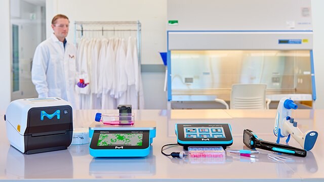 Five instruments sitting on a laboratory bench with a scientist in a lab coat walking past in the background.