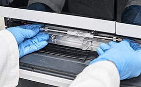 Ein Wissenschaftler in einem Labor, der blaue Handschuhe tr&auml;gt, setzt eine HPLC-S&auml;ule in ein Chromatographieger&auml;t f&uuml;r analytische Chromatographie ein.