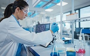 Ein Wissenschaftler in einem Labor, der einen wei&szlig;en Laborkittel, eine Schutzbrille und blaue Handschuhe tr&auml;gt, arbeitet mit analytischen Reagenzien und benutzt eine Pipette, um eine Fl&uuml;ssigkeit in einen Kolben zu transferieren. Gleichzeitig stehen auf dem Tisch verschiedene bunte L&ouml;sungen und Laborger&auml;te.