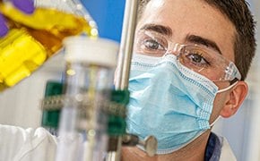 Ein Wissenschaftler in einem Labor, der einen wei&szlig;en Laborkittel, eine Schutzbrille, einen Mundschutz und lila Handschuhe tr&auml;gt, arbeitet mit chemisch-biologischen Werkzeugen. Er gie&szlig;t vorsichtig eine gelbe Fl&uuml;ssigkeit aus einem Becherglas in eine spezielle Glasapparatur. Der Hintergrund deutet auf eine gut ausgestattete Laborumgebung mit Schwerpunkt auf chemisch-biologischer Forschung hin.