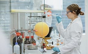 Eine Wissenschaftlerin im Laborkittel und mit Schutzbrille arbeitet in einem Labor, h&auml;lt eine Pipette und gibt vorsichtig eine Substanz in ein Reaktionsgef&auml;&szlig;. Der Labortisch ist voll mit verschiedenen Flaschen und Beh&auml;ltern mit chemischen Reagenzien sowie mit Glasgef&auml;&szlig;en und Ger&auml;ten f&uuml;r die chemische Synthese. Das Bild zeigt die pr&auml;zise und methodische Arbeit im Umgang mit chemischen Bausteinen, die f&uuml;r die Entwicklung neuer Verbindungen und die Durchf&uuml;hrung modernster Forschung unerl&auml;sslich sind.