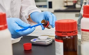 Ein Wissenschaftler in einem Labor mit blauen Handschuhen hantiert mit einer braunen Flasche mit chemischen Reagenzien von SigmaAldrich™, w&auml;hrend auf dem Arbeitstisch verschiedene andere Flaschen und Laborger&auml;te zu sehen sind, die auf eine aktive Forschungsumgebung hindeuten.