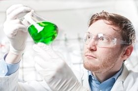 The image shows a scientist wearing safety goggles and gloves, carefully examining a flask containing a bright green liquid. The scientist appears to be deeply focused, possibly analyzing the solution or conducting an experiment. The laboratory background is softly blurred, directing attention to the scientist and the vivid color of the liquid in the flask, highlighting the precision and concentration required in scientific research.