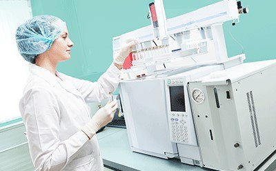 > Female scientific researcher with lab coat, gloves and hair protection placing vial flask in Gas Chromatography instrument