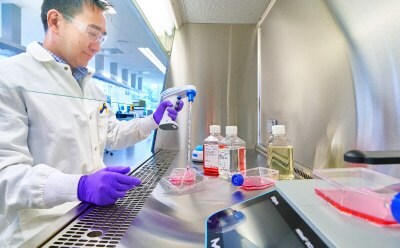 Male scientist sitting at the tissue culture hood holding a handheld pipette gun pipetting red media into a six well cell culture plate. Three media bottles are in the background and a T flask with media is in the foreground on a Millicell® DCI.