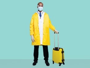 A person wearing a yellow raincoat over a white lab coat, black pants, and black shoes, standing next to a yellow suitcase with an "M" logo on it. 