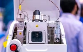 A mass spectrometer sits in focus in the center of the photo. Behind and to the right stands a scientist with his back turned