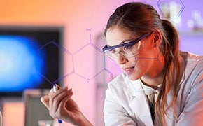 Brown-haired female scientist wearing white lab coat and safety goggles writes a chemical structure on glass screen. Computer screen and lab setting are visible in background.