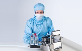 Five scientists wearing white lab coats, safety goggles and blue nitrile gloves work at different stations within the lab operating chemistry analytical instruments and filling glass beakers and vials. 