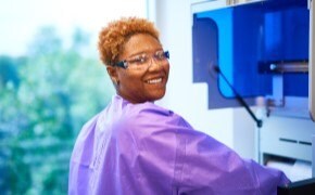 Female scientist in purple lab coat and safety goggles in molecular lab smiles into camera as she works with instrument preparing biosafety testing samples.