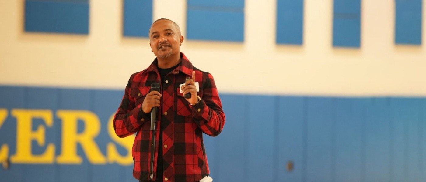 Abdi Wege wears a red flannel and holds a microphone in his right hand.