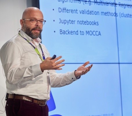 Daniel Kuhn gives a presentation wearing a white shirt. He uses his hands to gesture. Behind him a portion of a slide is visible. 