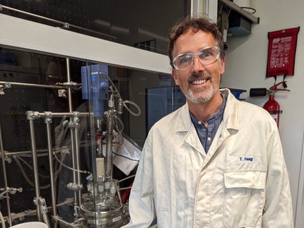 Haag stands in the lab in a lab coat and wearing safety glasses.