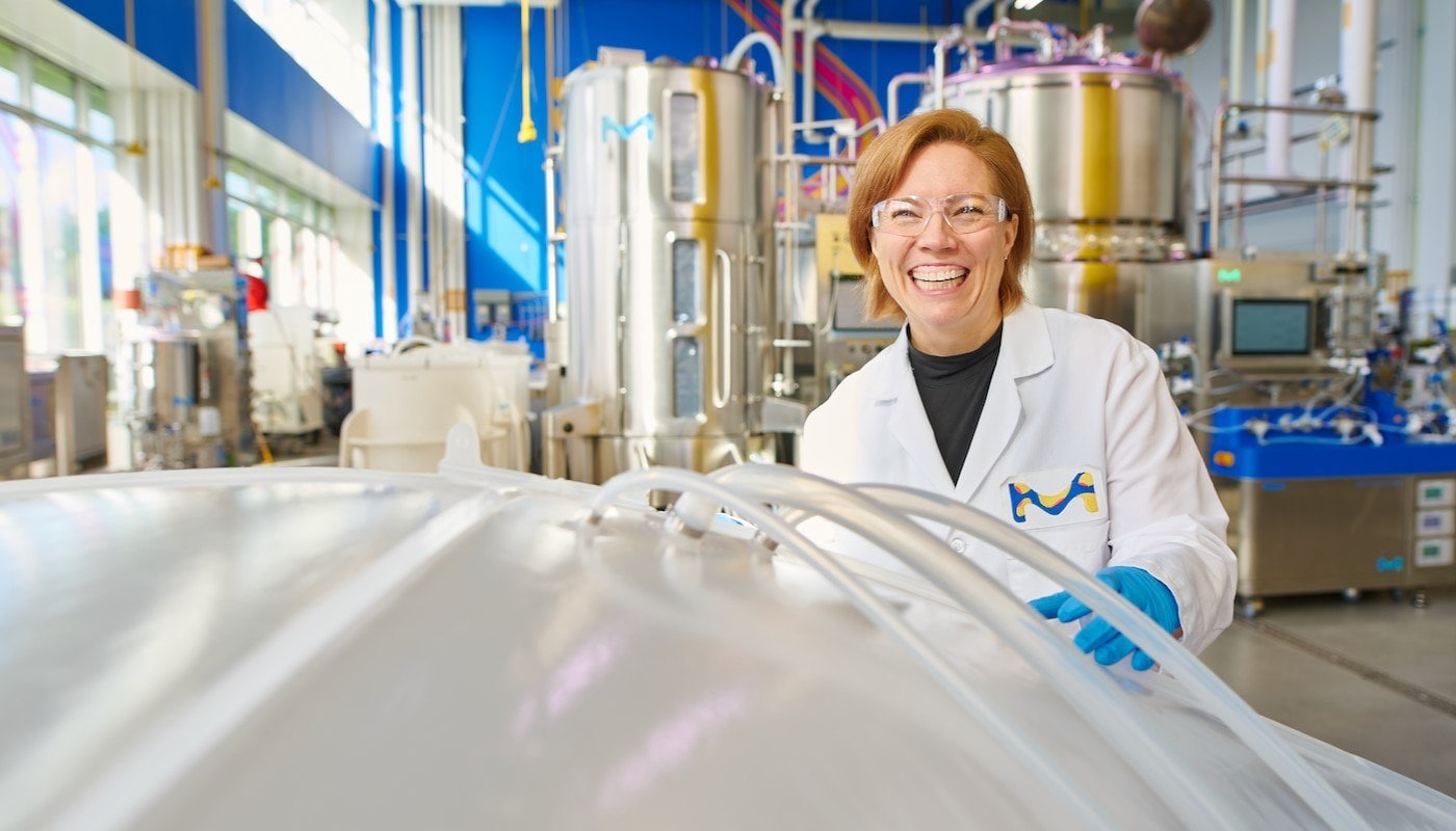 Shannon wears a lab coat, blue gloves and safety glasses and stands in an M Lab — a non-GMP biomanufacturing setting. She is smiling and holding onto a bin bag, which is situated in the lower left corner.