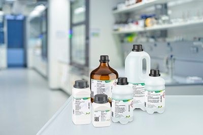 A variety of laboratory Emsure solvent bottles, labeled with product information, placed on a white counter in a modern laboratory setting. The bottles vary in size and shape, with both amber and white bottles visible, representing different chemical solvents typically used in analytical and research labs. The background features lab equipment and shelves