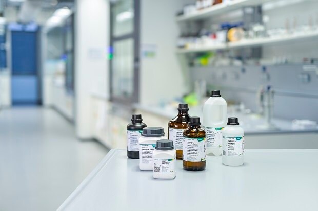 A variety of laboratory Emsure solvent bottles, labeled with product information, placed on a white counter in a modern laboratory setting. The bottles vary in size and shape, with both amber and white bottles visible, representing different chemical solvents typically used in analytical and research labs. The background features lab equipment and shelves