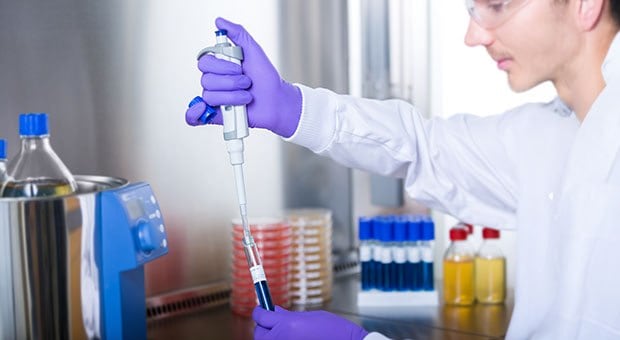 A lab technician in a white lab coat and purple gloves using a pipette to transfer liquid from a tube, with various ReadyTube® media in tubes and bottles for isolating and enumerating microorganisms in food visible in the background.