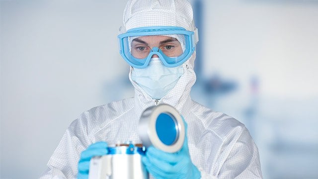 A person in a cleanroom suit and gloves is holding a metallic container with a blue lid. The background is a blurred laboratory setting, emphasizing a sterile and controlled environment.