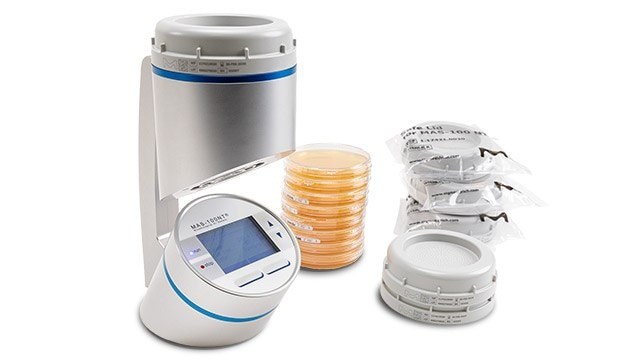 An advanced laboratory device is displayed, featuring a digital interface and a cylindrical chamber at the top. Next to it are several petri dishes containing orange media, along with sealed packages of additional laboratory supplies.